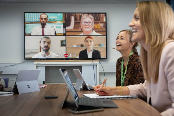 Two women sat at a meeting desk, withe people join the meeting via a screen on the back wall.